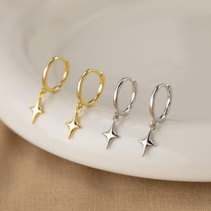 Puede incluir: Cuatro pendientes de aro con colgantes de estrella. Dos son dorados y dos plateados. Los pendientes se muestran sobre una superficie blanca y curvada, con un fondo de tono neutro.