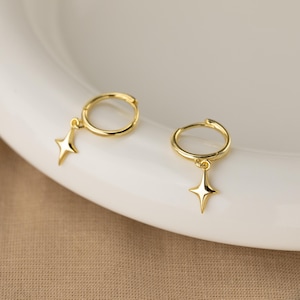 Puede incluir: Un par de pendientes de aro dorados. Cada pendiente presenta un peque&ntilde;o dije de estrella de cuatro puntas colgando del aro. Los pendientes se exhiben sobre una superficie blanca, con un fondo de tono neutro.