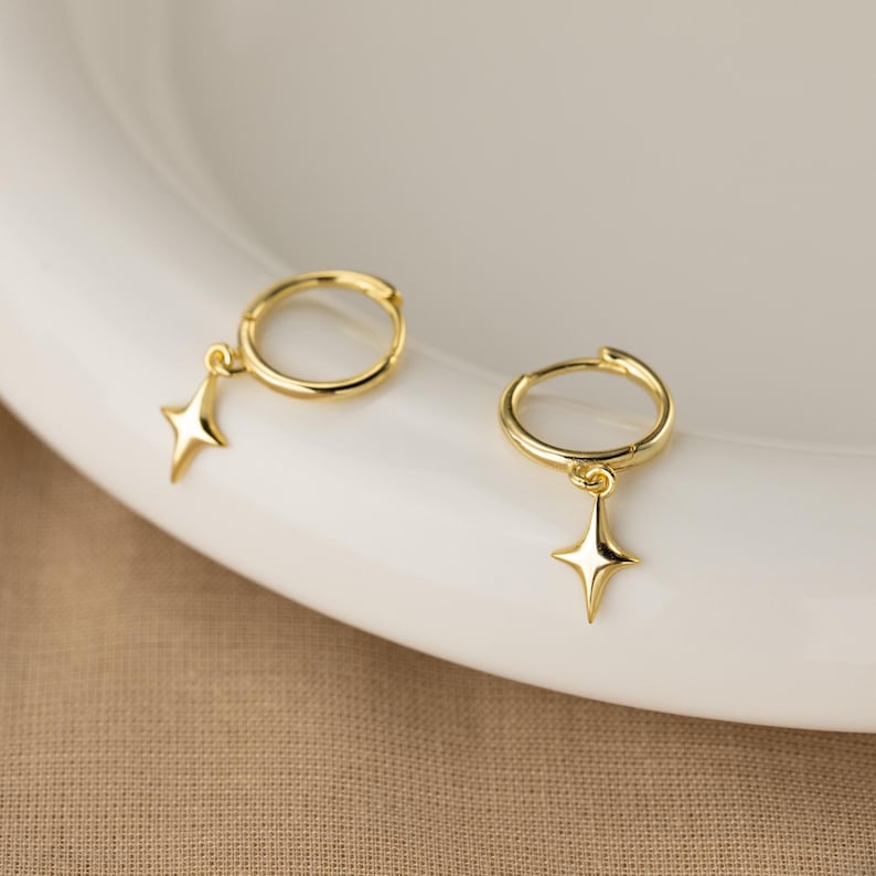 Puede incluir: Un par de pendientes de aro dorados. Cada pendiente presenta un peque&ntilde;o dije de estrella de cuatro puntas colgando del aro. Los pendientes se exhiben sobre una superficie blanca, con un fondo de tono neutro.