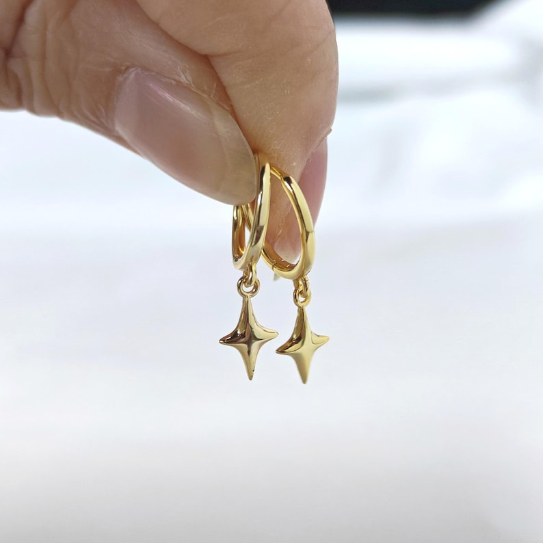 Puede incluir: Un par de pendientes de aro dorados con colgantes de estrella. Los pendientes se sostienen sobre un fondo blanco. Los colgantes de estrella tienen cuatro puntas y son de color dorado brillante.