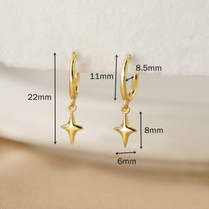 Puede incluir: Pendientes de aro dorados con colgantes en forma de estrella. Los aros tienen 11 mm de di&aacute;metro, y los colgantes miden 8 mm de largo y 6 mm de ancho. Los pendientes miden 22 mm de largo en total. Se muestran sobre una superficie blanca.