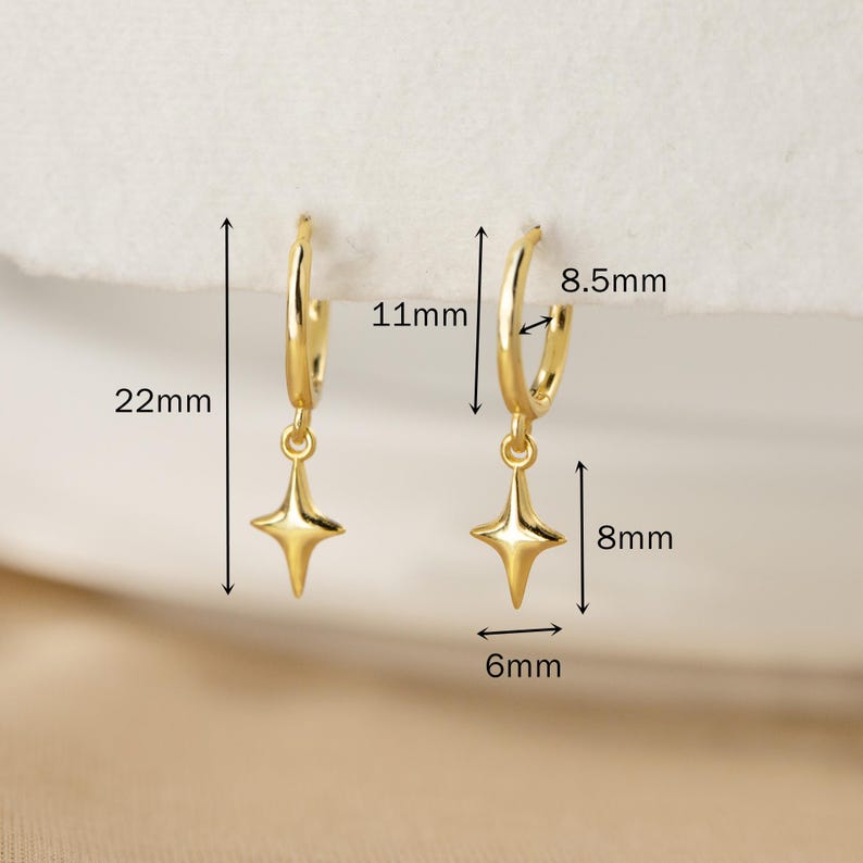 Puede incluir: Pendientes de aro dorados con colgantes en forma de estrella. Los aros tienen 11 mm de di&aacute;metro, y los colgantes miden 8 mm de largo y 6 mm de ancho. Los pendientes miden 22 mm de largo en total. Se muestran sobre una superficie blanca.