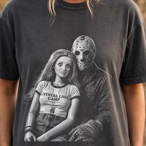 Peut inclure: Un t-shirt gris anthracite &agrave; manches courtes avec une impression graphique repr&eacute;sentant une poup&eacute;e aux longs cheveux portant un t-shirt &agrave; col contrast&eacute; marqu&eacute; CRYSTAL LAKE CAMP, assise &agrave; c&ocirc;t&eacute; d'une silhouette portant un masque de hockey.