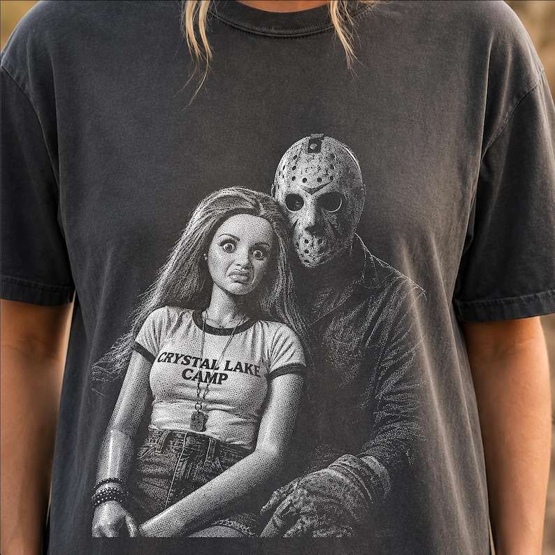 Peut inclure: Un t-shirt gris anthracite &agrave; manches courtes avec une impression graphique repr&eacute;sentant une poup&eacute;e aux longs cheveux portant un t-shirt &agrave; col contrast&eacute; marqu&eacute; CRYSTAL LAKE CAMP, assise &agrave; c&ocirc;t&eacute; d'une silhouette portant un masque de hockey.