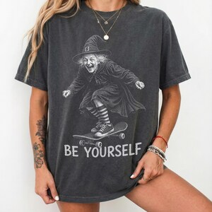 May include: A charcoal gray short-sleeve t-shirt featuring a graphic illustration of a witch wearing a pointed hat and striped socks while riding a skateboard. The text "BE YOURSELF" is printed in white capital letters below the illustration.