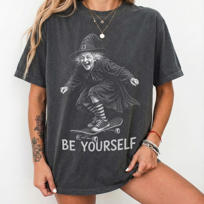 May include: A charcoal gray short-sleeve t-shirt featuring a graphic illustration of a witch wearing a pointed hat and striped socks while riding a skateboard. The text "BE YOURSELF" is printed in white capital letters below the illustration.