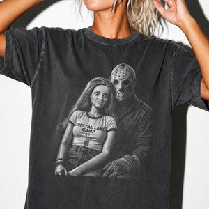 Peut inclure: Un t-shirt gris anthracite &agrave; manches courtes avec une impression graphique repr&eacute;sentant une poup&eacute;e aux longs cheveux portant un t-shirt marqu&eacute; CRYSTAL LAKE CAMP, &agrave; c&ocirc;t&eacute; d'une silhouette portant un masque de hockey. Effet d&eacute;lav&eacute; vintage.