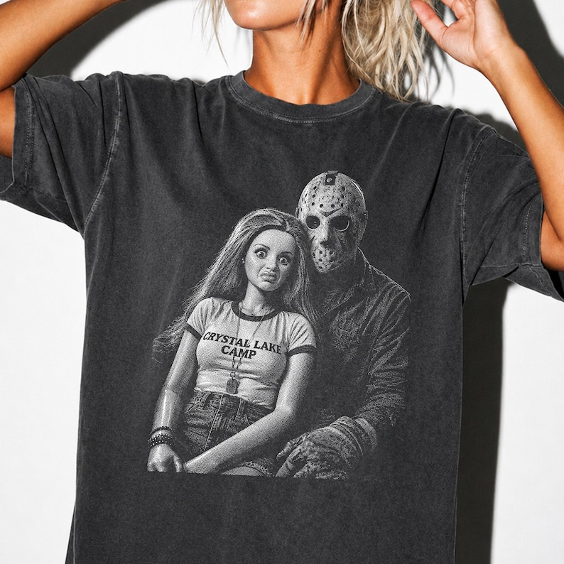 Peut inclure: Un t-shirt gris anthracite &agrave; manches courtes avec une impression graphique repr&eacute;sentant une poup&eacute;e aux longs cheveux portant un t-shirt marqu&eacute; CRYSTAL LAKE CAMP, &agrave; c&ocirc;t&eacute; d'une silhouette portant un masque de hockey. Effet d&eacute;lav&eacute; vintage.