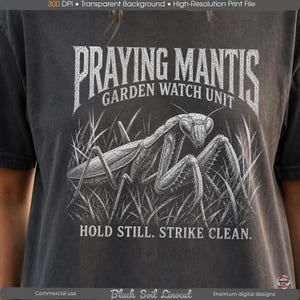 May include: A charcoal gray t-shirt featuring a white graphic of a praying mantis in tall grass. The text reads PRAYING MANTIS GARDEN WATCH UNIT and HOLD STILL. STRIKE CLEAN. The design has a distressed, textured appearance with a vintage aesthetic.