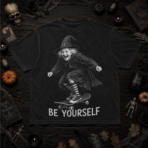 May include: A black t-shirt featuring a graphic of a witch wearing a pointed hat and striped socks while riding a skateboard. The witch has a wide, joyful expression. The text "BE YOURSELF" is printed in white capital letters below the graphic.
