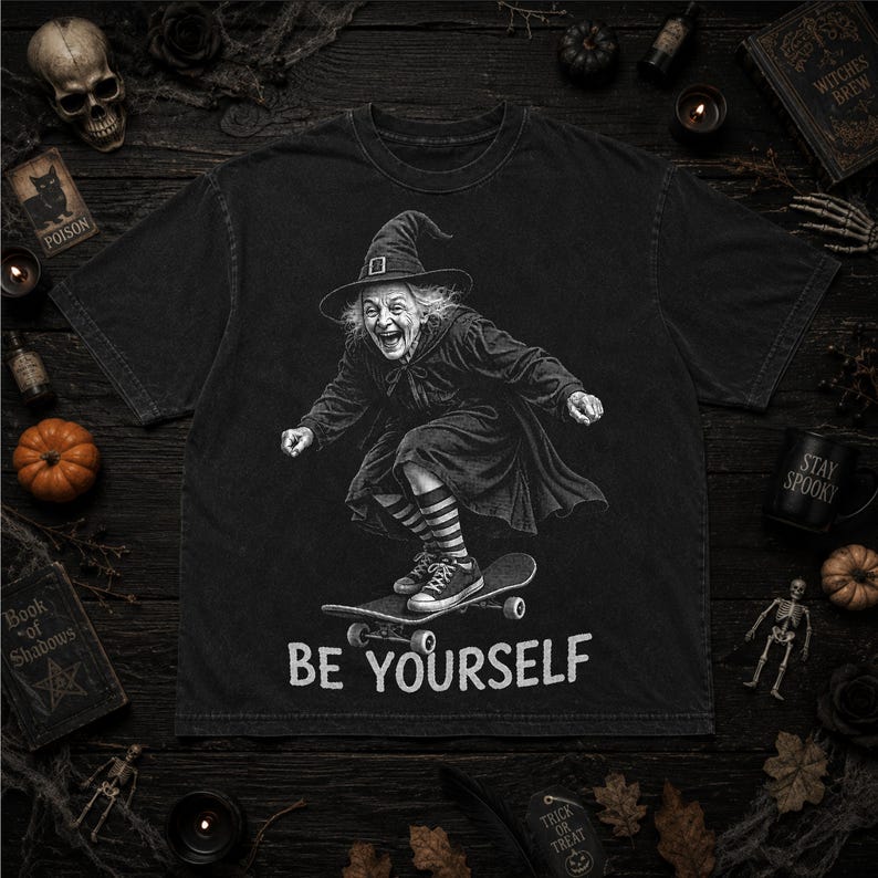 May include: A black t-shirt featuring a graphic of a witch wearing a pointed hat and striped socks while riding a skateboard. The witch has a wide, joyful expression. The text "BE YOURSELF" is printed in white capital letters below the graphic.