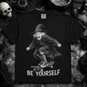May include: A black short-sleeve t-shirt featuring a graphic of a witch wearing a pointed hat and striped socks while riding a skateboard. Below the graphic, the text reads BE YOURSELF in a distressed white font. The shirt has a crew neckline.