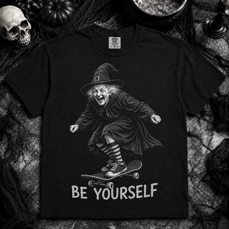May include: A black short-sleeve t-shirt featuring a graphic of a witch wearing a pointed hat and striped socks while riding a skateboard. Below the graphic, the text reads BE YOURSELF in a distressed white font. The shirt has a crew neckline.