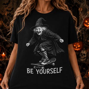 May include: A black t-shirt featuring a white graphic of an elderly person wearing a witch hat and a cloak while riding a skateboard. The person wears striped socks and sneakers. The text "BE YOURSELF" is printed in white below the graphic.