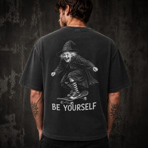 May include: A charcoal gray short-sleeve t-shirt featuring a graphic on the back of a witch wearing a pointed hat and striped socks while riding a skateboard. Below the graphic, the text BE YOURSELF is printed in a bold, white, sans-serif font.