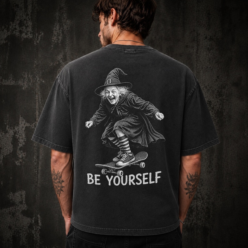May include: A charcoal gray short-sleeve t-shirt featuring a graphic on the back of a witch wearing a pointed hat and striped socks while riding a skateboard. Below the graphic, the text BE YOURSELF is printed in a bold, white, sans-serif font.