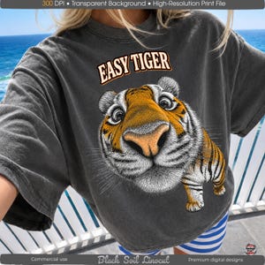 Może przedstawiać: Ciemnoszara koszulka z grafiką przedstawiającą twarz tygrysa i napisem "EASY TIGER". Tygrys ma pomarańczowe i czarne paski, duży nos i duże oczy. Koszulka ma luźny krój.