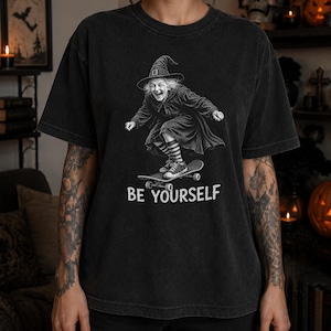 May include: A charcoal black short-sleeve t-shirt featuring a graphic of a witch wearing a pointed hat, a long dress, and striped socks while riding a skateboard. The text "BE YOURSELF" is printed in white capital letters below the graphic.