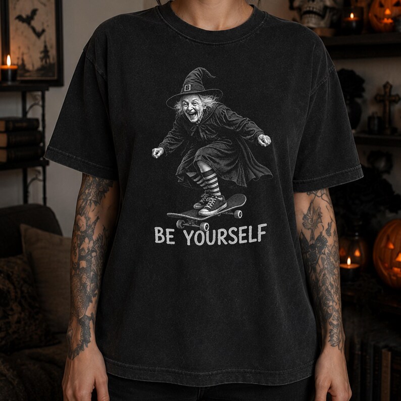 May include: A charcoal black short-sleeve t-shirt featuring a graphic of a witch wearing a pointed hat, a long dress, and striped socks while riding a skateboard. The text "BE YOURSELF" is printed in white capital letters below the graphic.