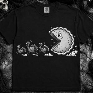 May include: A black short-sleeve t-shirt featuring a white graphic print of three turkeys running away from a large, circular, pie-shaped character with a wide-open mouth. The shirt has a crew neckline and a Comfort Colors brand label at the inner neck.