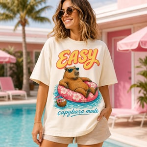 Op de afbeelding: Crèmekleurig T-shirt met het woord "EASY" in gele en rode letters. Het shirt heeft een cartoon capibara met een zonnebril die op een roze zwemband drijft. De tekst "capybara mode" staat eronder.