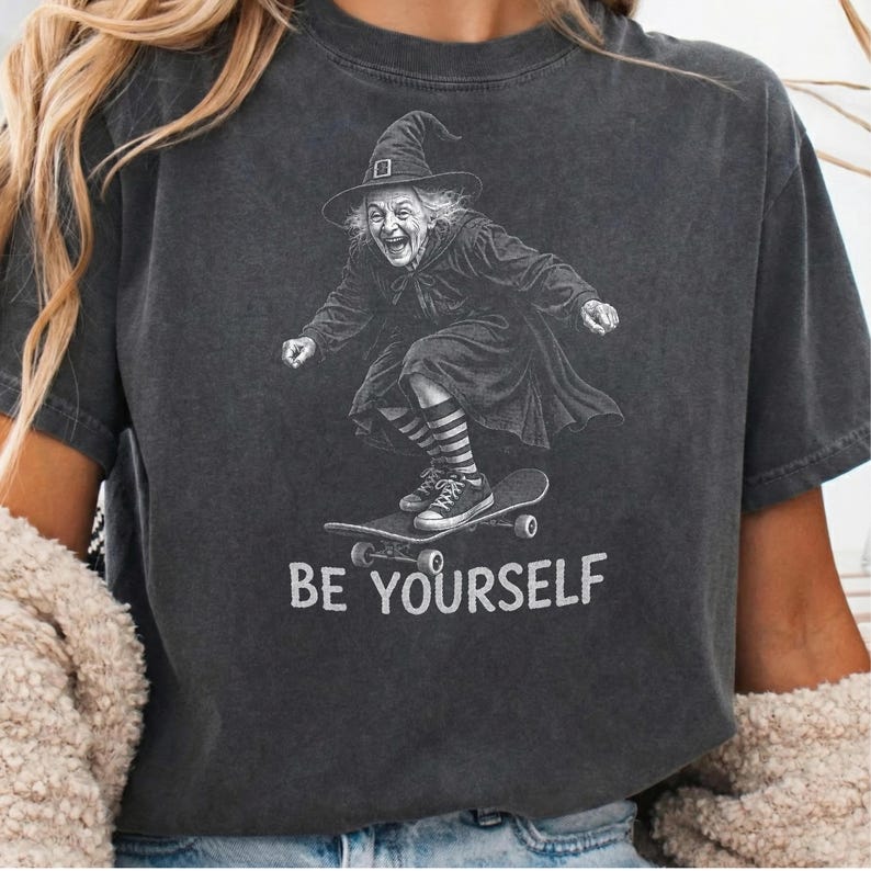 May include: A charcoal gray short-sleeve t-shirt featuring a graphic of a witch wearing a pointed hat and striped socks while riding a skateboard. Below the graphic, the text BE YOURSELF is printed in a bold, white, sans-serif font.