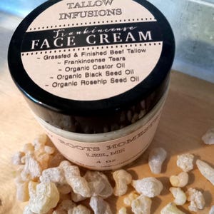 May include: A black jar with a brown label that reads "Frankincense Face Cream" and lists the ingredients: Grassfed & Finished Beef Tallow, Frankincense Tears, Organic Castor Oil, Organic Black Seed Oil, Organic Rosehip Seed Oil. The jar is sitting on a wooden surface with a pile of frankincense resin in front of it.