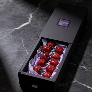 May include: A black box with a drawer, containing red, cherry-shaped candies with green stems, nestled on purple tissue paper. The box has a small purple label with white text. The candies are arranged in rows.