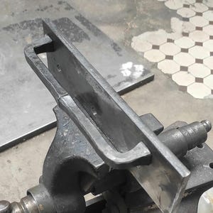 Könnte beinhalten: Ein Metallobjekt, das in einem Schraubstock gehalten wird. Das Objekt hat eine rechteckige Form mit einem Griff. Der Schraubstock und das Objekt sind beide dunkelgrau. Der Hintergrund ist eine hellgraue Oberfläche und ein Fliesenboden.