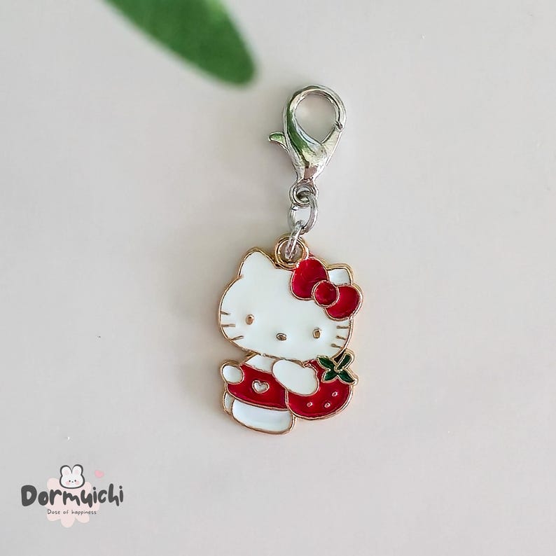 May include: A white and red enamel charm featuring a cartoon cat with a red bow and a strawberry. The charm has a silver-colored clasp and is a cute accessory. The charm is a fun, colorful item.