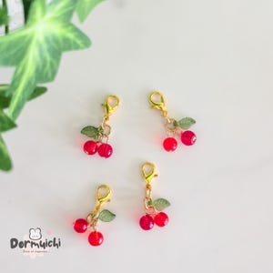 May include: Four cherry-themed charms with gold-colored clasps. Each charm features two red, translucent cherry beads and two green leaf beads. The charms are arranged on a white surface with green foliage in the background.