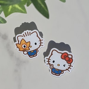 May include: Two white Hello Kitty stickers. One sticker has a red bow, and the other has a yellow flower. Both stickers have a blue dress and are on a white background.