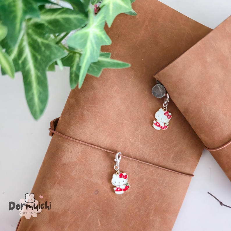 May include: Two brown leather journals with Hello Kitty charms attached. The charms are white with red accents and a red bow. The journals are closed and tied with a brown cord. Green leaves are in the background.
