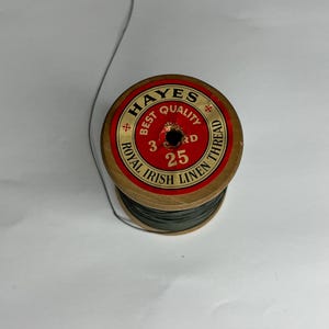 Puede incluir: Una bobina de hilo verde oscuro con una bobina de madera. La etiqueta dice "Hayes Best Quality Royal Irish Linen Thread 3 Cord 25".