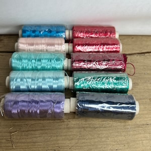 May include: Eight spools of thread in various colours, including blue, pink, teal, green, purple, and navy. The spools are wrapped in clear plastic and have white cardboard cores.