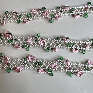 Op de afbeelding: Close-up van drie rijen decoratieve sierband. De sierband heeft een witte, open geweven basis met roze satijnen rozen en groene accenten. De sierband wordt waarschijnlijk gebruikt voor naai- of knutselprojecten.