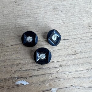 May include: Three black buttons, each with a central, clear rhinestone. The buttons feature a geometric design, with one round and two square-shaped. They are set against a weathered, light brown wooden surface.