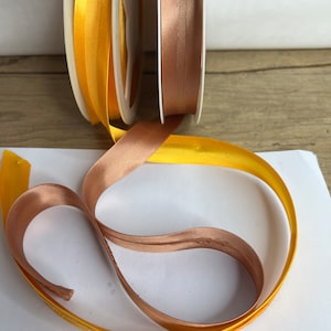 May include: Two spools of ribbon, one gold and one brown, are unwound and laid out on a white surface. The ribbon is satin and has a subtle sheen.
