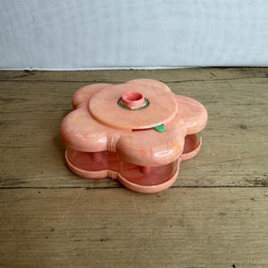 May include: A vintage, flower-shaped container in shades of pink. The container has a central opening and several compartments. The design includes a green button and a red line. The container is made of plastic and sits on a wooden surface.