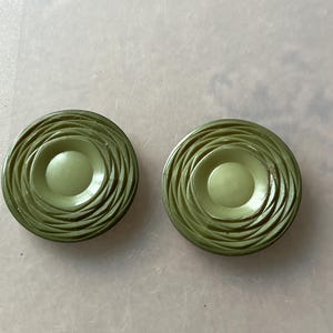 May include: Two olive green buttons with a concentric spiral design. Each button has a raised center and a series of curved ridges radiating outwards. The buttons are made of a solid, opaque material.
