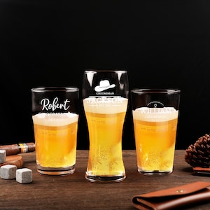 May include: Three clear beer glasses filled with beer, each with a different design and text. The glasses are on a wooden surface with a black background. One glass says "Robert", another says "Jackson", and the third says "William".