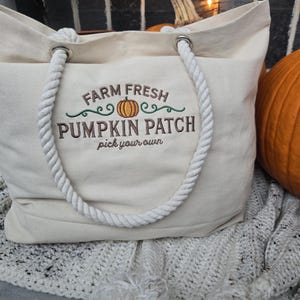 May include: A white canvas tote bag with a rope handle. The bag has an embroidered design that says "Farm Fresh Pumpkin Patch pick your own".