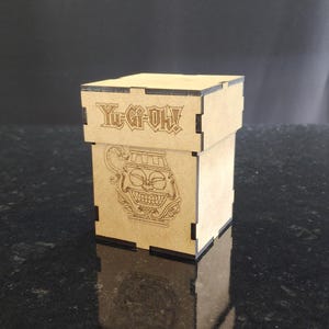 May include: A wooden box with a brown finish and black trim. The box has a cutout design on the front with the text "Yu-Gi-Oh!" and a cartoon image of a monster.