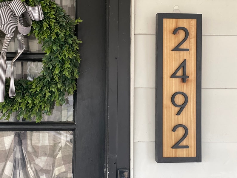 May include: A rectangular wooden address sign with the numbers 2492 in black. The sign has a light wood center and a dark gray frame. The sign is mounted on a white wall. A green wreath with a striped bow is visible in the background.