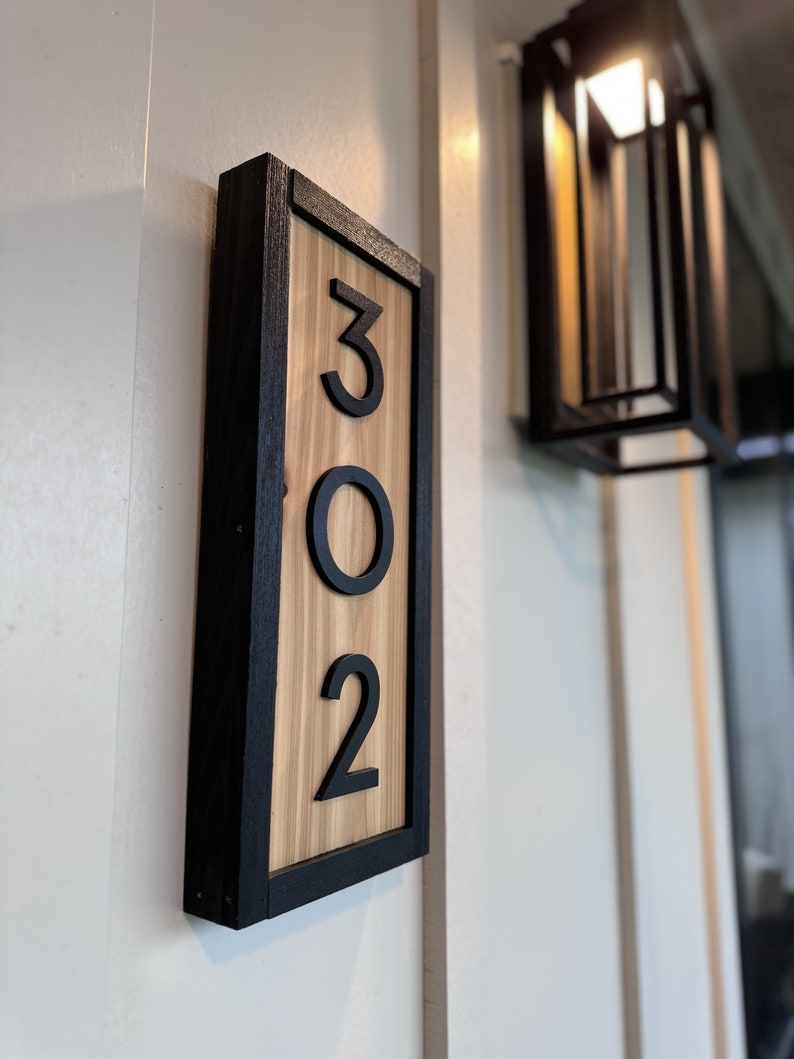 May include: A rectangular wooden address sign with the numbers 302. The numbers are black and raised against a light wood grain background. The sign is framed in black and mounted on a white wall.