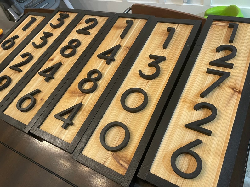 May include: Wooden address plaques with black numbers. Each plaque features a natural wood grain background framed by a black border. The numbers are in a bold, modern font, providing a clear and stylish display of house numbers.