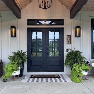 May include: Exterior view of a home's entrance featuring black double doors, a welcome mat, and a striped rug. The porch is decorated with potted plants and wall-mounted lights. A black lantern hangs from the wooden ceiling.