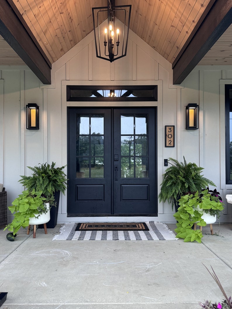 May include: Exterior view of a home's entrance featuring black double doors, a welcome mat, and a striped rug. The porch is decorated with potted plants and wall-mounted lights. A black lantern hangs from the wooden ceiling.