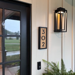 May include: A wooden address sign with the numbers "302" in a black frame is mounted on a cream-colored wall. A black outdoor light fixture and a doorbell are also visible. Green ferns are in the foreground.
