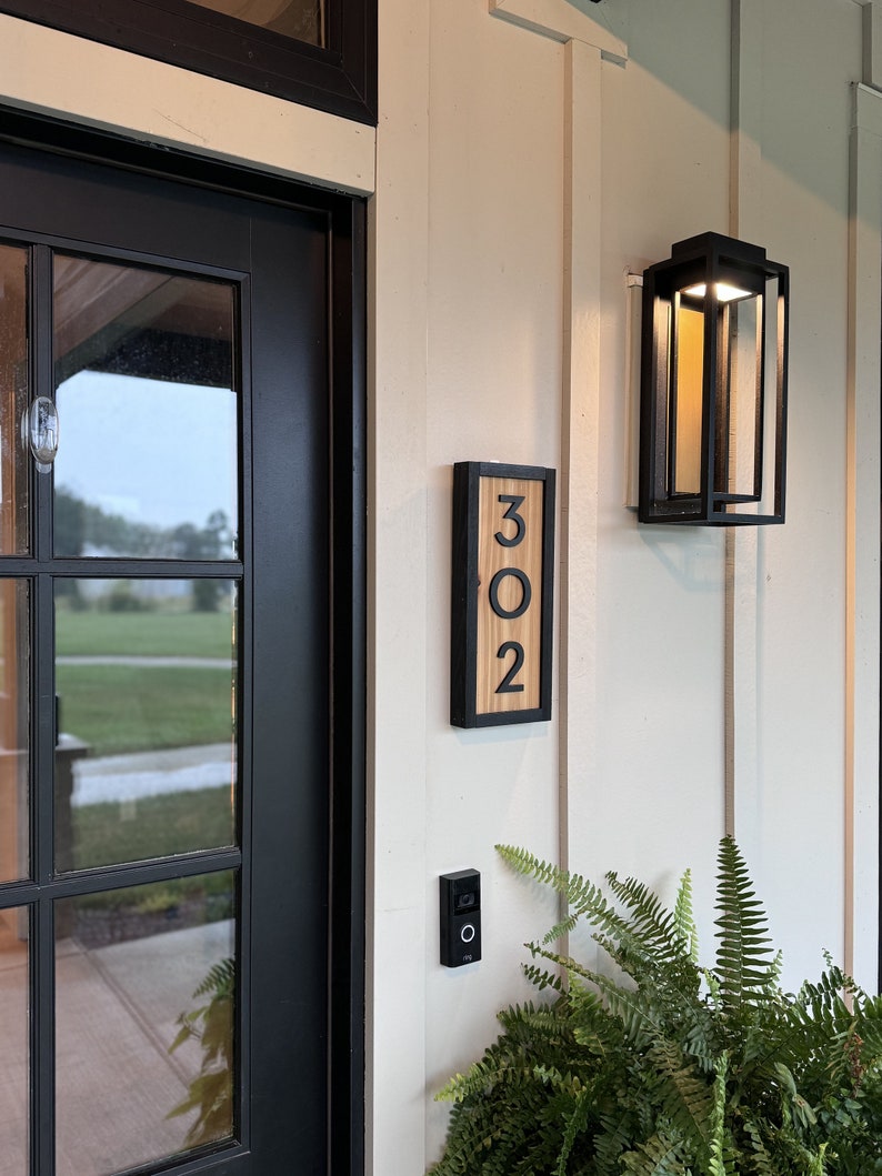 May include: A wooden address sign with the numbers "302" in a black frame is mounted on a cream-colored wall. A black outdoor light fixture and a doorbell are also visible. Green ferns are in the foreground.